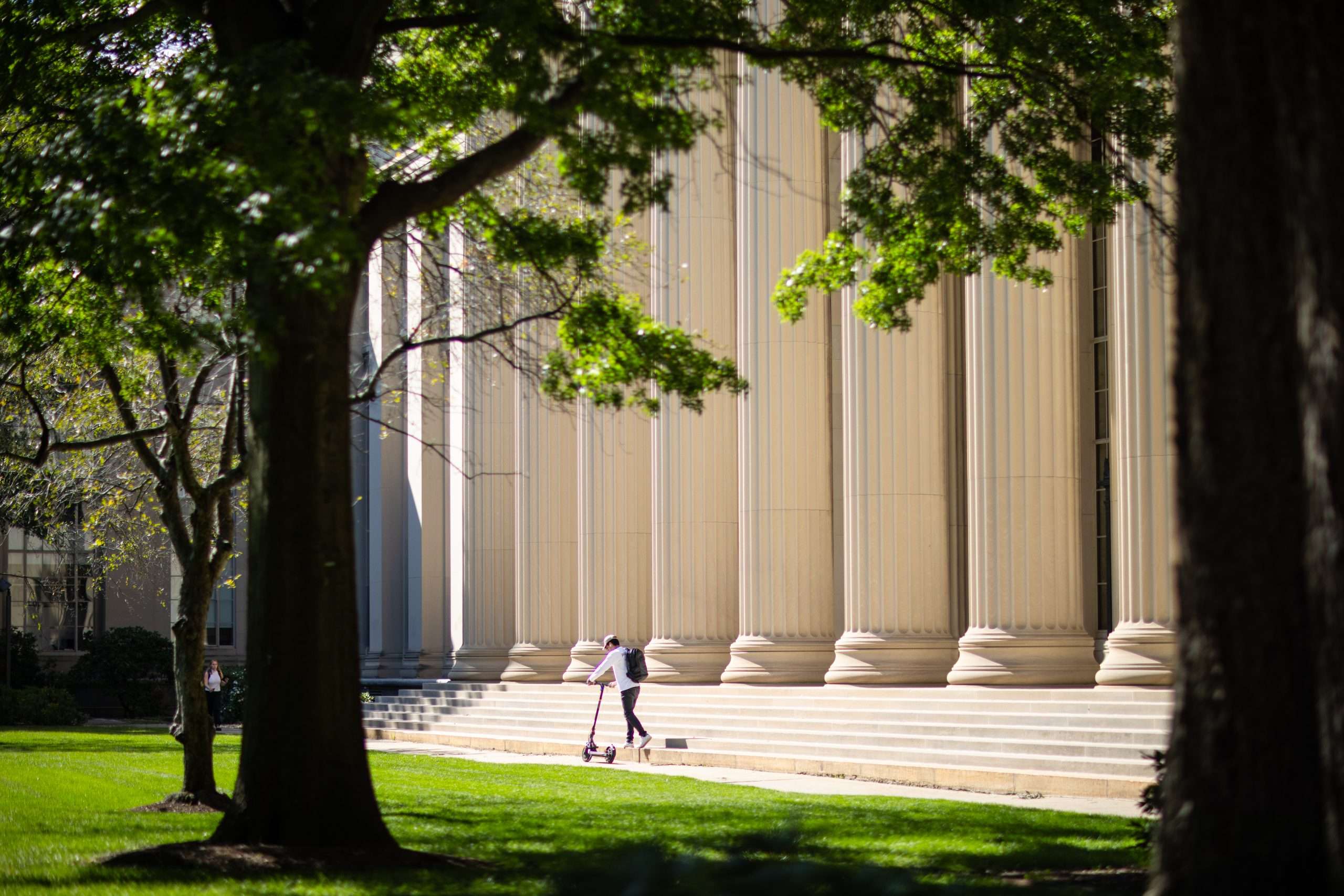 A student walks across the steps of a columned building at MIT with a scooter.