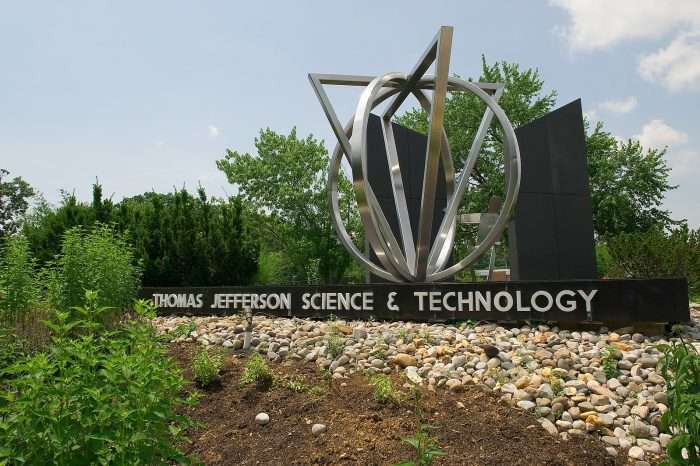 A metal, circular sculpture with the words "Thomas Jefferson Science & Technology" inscribed beneath it stands behind a dirt patch at Thomas Jefferson High School for Science and Technology.